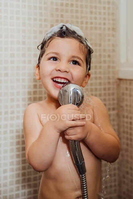 Niño sonriente con espuma en la cabeza de pie en el baño con ducha y cantando mientras mira hacia otro lado - foto de stock