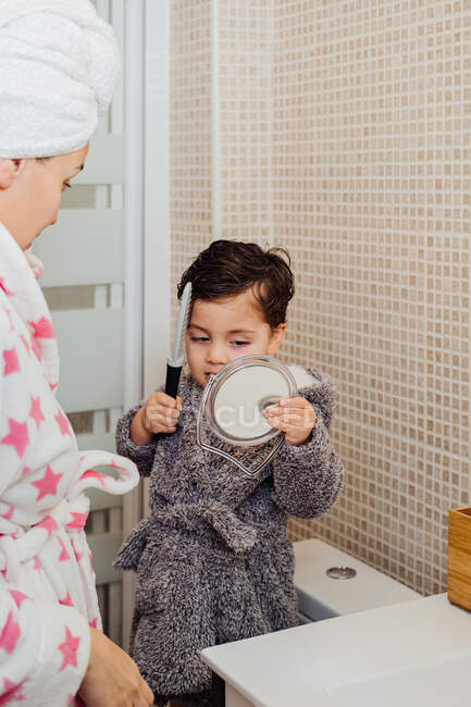 Красивый ребенок в халате стоит в яркой ванной комнате вместе с матерью и расчесывая мокрые волосы после душа, глядя в зеркало — стоковое фото