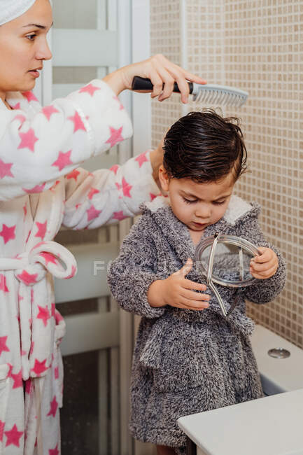 Side view of mother wearing bathrobe and towel turban combing wet hair of little kid while standing in modern bathroom — Stock Photo