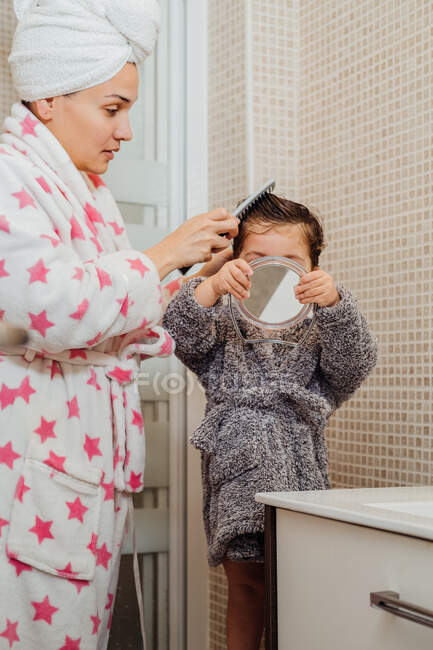 Vista laterale della madre che indossa accappatoio e asciugamano turbante pettinare i capelli bagnati del bambino mentre in piedi in bagno moderno — Foto stock