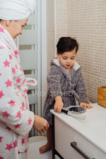 Criança adorável em roupão de banho em pé no banheiro brilhante, juntamente com a mãe e pentear o cabelo molhado após o chuveiro enquanto olha no espelho — Fotografia de Stock