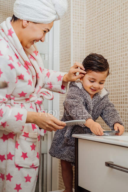 Vista lateral de la madre con albornoz y turbante toalla peinando el cabello mojado de un niño pequeño mientras está de pie en el baño moderno - foto de stock