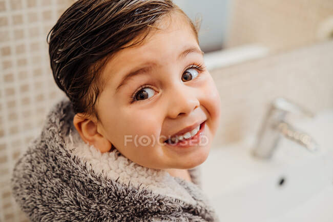 Сверху веселый маленький ребенок с мокрыми волосами в мягком халате стоит возле раковины в ванной комнате и смотрит в камеру — стоковое фото