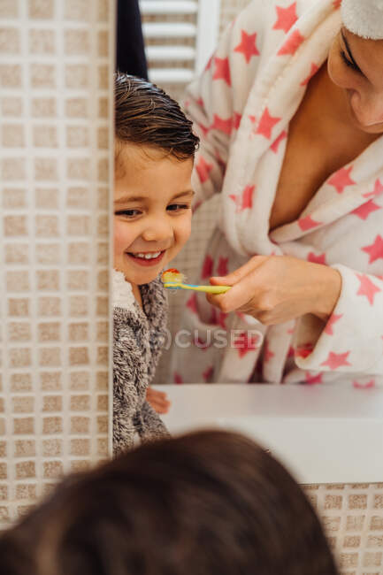 Petit garçon mignon en peignoir et mère souriante en serviette turban debout dans la salle de bain et brossant les dents — Photo de stock