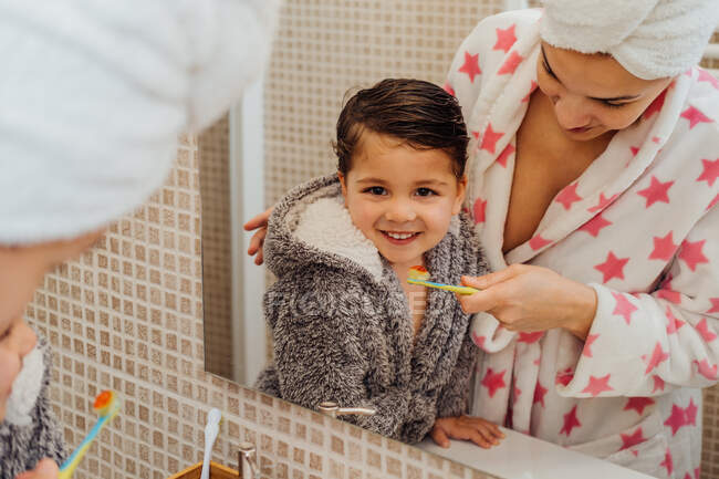 Симпатичний маленький хлопчик в халаті і усміхнена мати в рушнику турбан, що стоїть у ванній кімнаті і чистить зуби — стокове фото