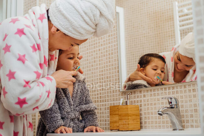 Cute little boy in bathrobe and smiling mother in towel turban standing in bathroom and brushing teeth — Stock Photo