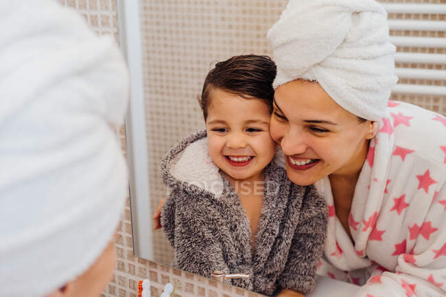 Cheerful woman with towel turban cuddling little child in bathrobe after taking shower and looking in the mirror — Stock Photo