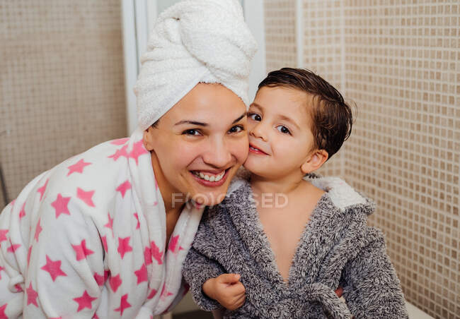 Donna allegra con asciugamano turbante coccolando bambino in accappatoio dopo aver fatto la doccia e guardando la fotocamera — Foto stock