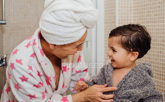 Fröhliche Frau mit Handtuch-Turban kuschelt kleines Kind im Bademantel, nachdem sie duscht und einander ansieht — Stockfoto