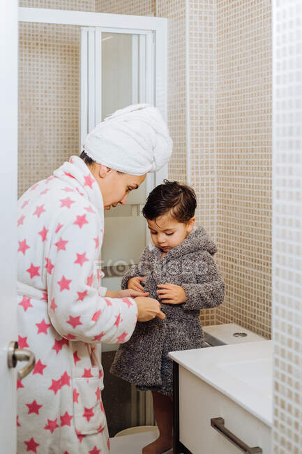 Cheerful woman with towel turban cuddling little child in bathrobe after taking shower — Stock Photo