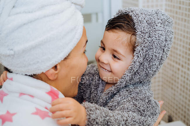 Вид ззаду на невпізнавану жінку з маленькою дитиною в халаті після прийняття душу і погляду один на одного — стокове фото