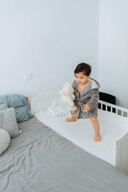 De cima de criança bonita com cabelo molhado em pé no berço no quarto enquanto brincava com brinquedo macio e se divertia durante o fim de semana — Fotografia de Stock