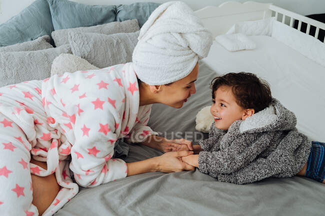 Mujer alegre en albornoz divirtiéndose con niño en la cama suave mirándose el uno al otro - foto de stock