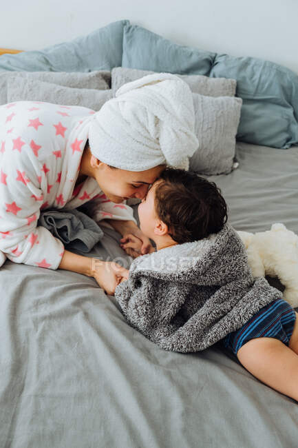 Сверху довольная женщина в халате веселится с маленьким мальчиком на мягкой кровати, смотрящим друг на друга — стоковое фото