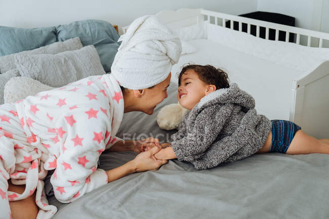 Femme gaie en peignoir s'amusant avec un petit garçon sur un lit doux en se regardant — Photo de stock