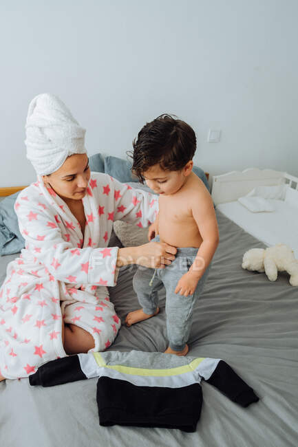 Mujer alegre en albornoz vestirse hijo pequeño mientras juegan juntos en casa mirándose el uno al otro - foto de stock