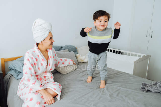 Femme joyeuse en peignoir habillant petit fils tout en jouant ensemble à la maison en se regardant — Photo de stock
