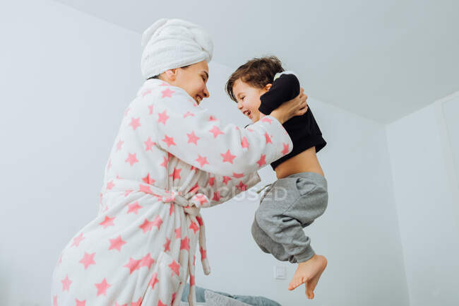 De baixo vista lateral da mulher alegre em roupão de banho jogando pequeno filho enquanto brincam juntos durante o fim de semana em casa olhando um para o outro — Fotografia de Stock