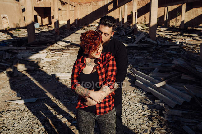 Positiva giovane coppia hipster con tatuaggi godendo del tempo insieme e abbracciandosi mentre in piedi contro la costruzione di pietra squallida nella giornata di sole — Foto stock