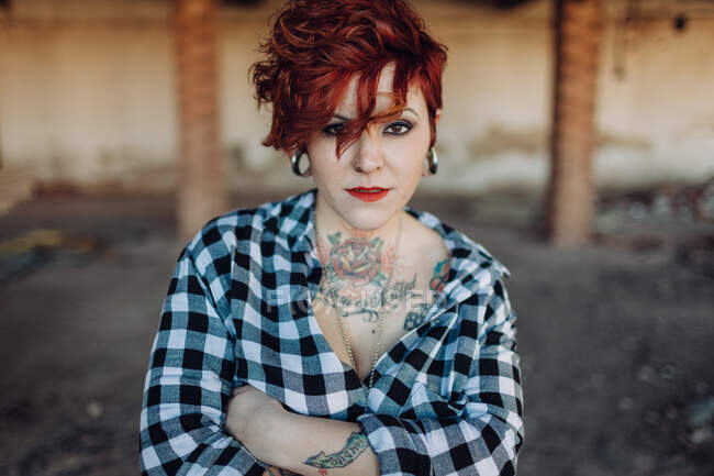 Mulher jovem e elegante independente com corte de cabelo na moda e tatuagens vestindo camisa xadrez casual olhando para a câmera enquanto está de pé contra o edifício gasto borrado — Fotografia de Stock