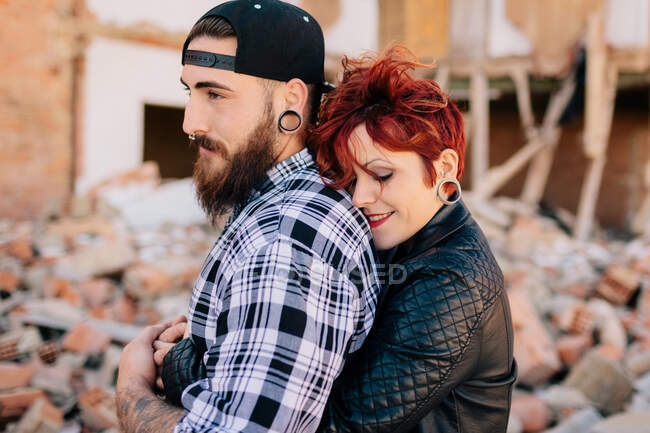 Positive young hipster couple with tattoos enjoying time together and hugging while standing against shabby stone construction in sunny day — Stock Photo