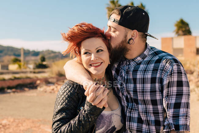 Positif jeune couple hipster avec des tatouages profiter du temps ensemble et étreindre tout en se tenant contre la construction en pierre minable dans la journée ensoleillée — Photo de stock