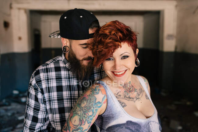 Positif jeune couple hipster avec des tatouages profiter du temps ensemble et étreindre tout en se tenant contre la construction en pierre minable dans la journée ensoleillée — Photo de stock