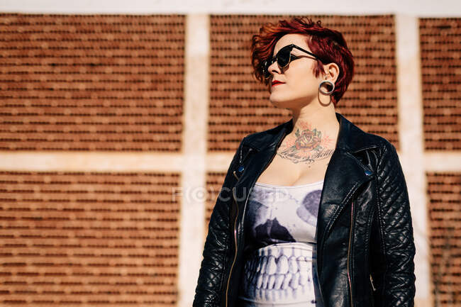 Mujer joven y elegante independiente con corte de pelo de moda y tatuajes con camisa a cuadros casual mirando a la cámara mientras está de pie contra el edificio borroso - foto de stock
