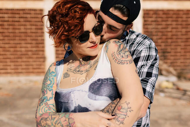 Positivo jovem casal hipster com tatuagens desfrutando de tempo juntos e abraçando enquanto de pé contra a construção de pedra gasto em dia ensolarado — Fotografia de Stock