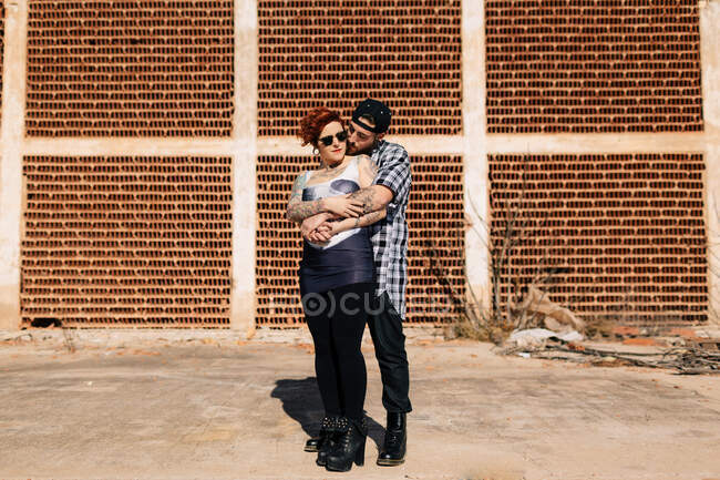 Позитивная молодая хипстерская пара с татуировками, наслаждающаяся временем вместе и обнимающаяся, стоя против обветшалого каменного строительства в солнечный день — стоковое фото