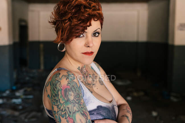Mujer joven y elegante independiente con corte de pelo de moda y tatuajes con camisa a cuadros casual mirando a la cámara mientras está de pie contra el edificio borroso - foto de stock