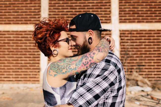 Позитивная молодая хипстерская пара с татуировками, наслаждающаяся временем вместе и обнимающаяся, стоя против обветшалого каменного строительства в солнечный день — стоковое фото