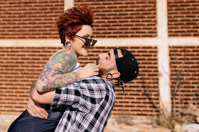 Side view of happy young man embracing hipster tattooed girlfriend while having fun during romantic date on city street near brick building — Stock Photo