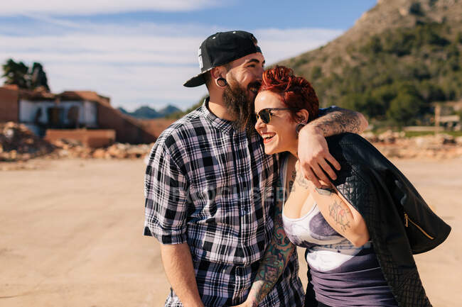 Positives junges Hipster-Paar mit Tätowierungen genießt die gemeinsame Zeit und umarmt sich, während es bei sonnigem Wetter vor schäbigen Steinbauten steht — Stockfoto