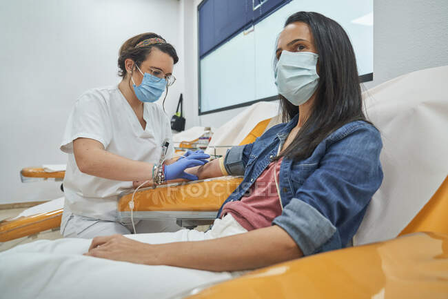 Бічний погляд на молоду жінку в захисній масці сидячи в медичному кріслі під час переливання крові в сучасній лікарні. — стокове фото
