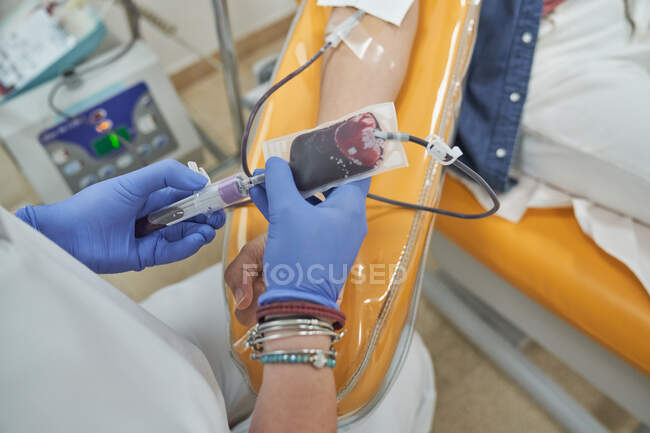 From above of crop female nurse in protective gloves with bag of blood in hand working with patient during procedure of blood donation in modern medical center — Stock Photo