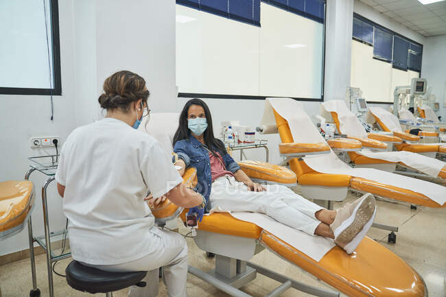 Вид сбоку молодой женщины в защитной маске, сидящей в медицинском кресле во время процедуры переливания крови в современной больнице — стоковое фото