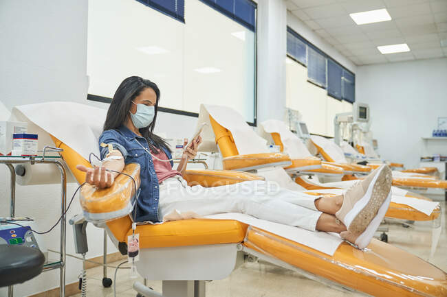 Вид з боку низького кута молодої жінки в захисній масці перегляду смартфона під час процедури переливання крові в лікарні — стокове фото