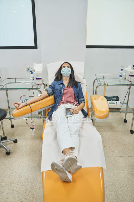 Von oben eine entspannte junge Frau in Schutzmaske mit Smartphone, die während der Blutspende im modernen Krankenhaus im Behandlungsstuhl sitzt — Stockfoto