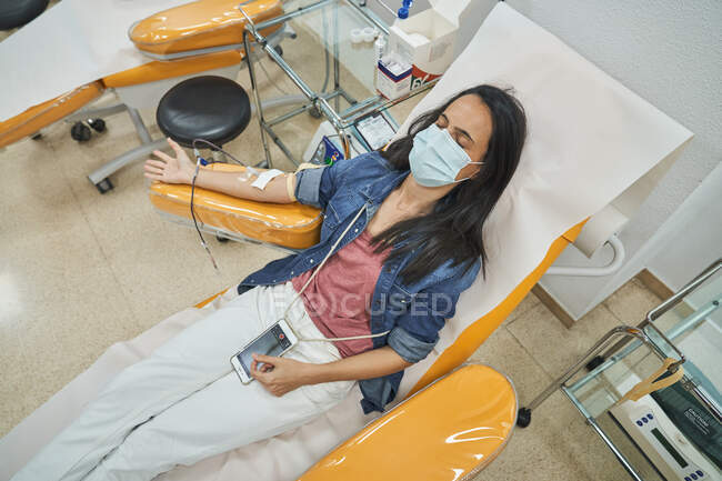 Desde arriba de mujer joven relajada en máscara protectora con teléfono inteligente sentado en silla médica durante la donación de sangre en el hospital moderno - foto de stock