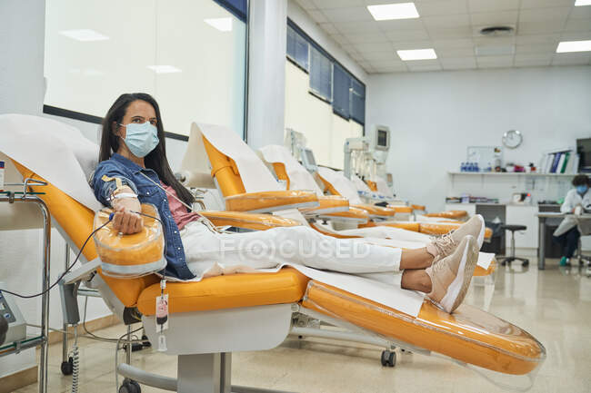 Vista laterale della giovane donna in maschera protettiva seduta sulla poltrona medica durante la procedura di trasfusione di sangue nell'ospedale contemporaneo — Foto stock