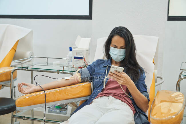 Нижчий кут зору молодої жінки в захисній масці, яка переглядає смартфон під час процедури переливання крові в лікарні. — стокове фото