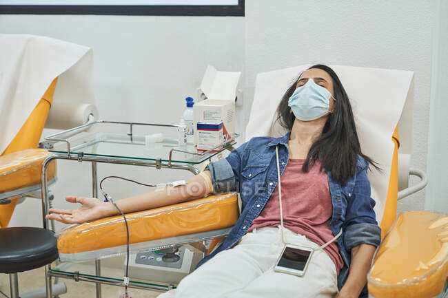 From above of relaxed young female in protective mask with smartphone sitting in medical chair during blood donation in modern hospital — Stock Photo
