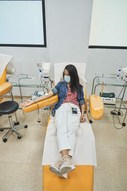 Mujer en máscara protectora sentada en sillón médico durante el procedimiento de transfusión de sangre en el hospital contemporáneo - foto de stock