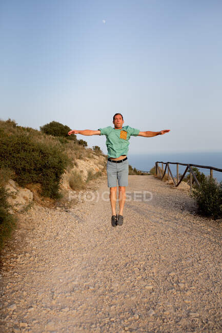 Full length young male in casual wear jumping with outstretched arms over sandy pathway leading along sea shore in summer evening — Stock Photo