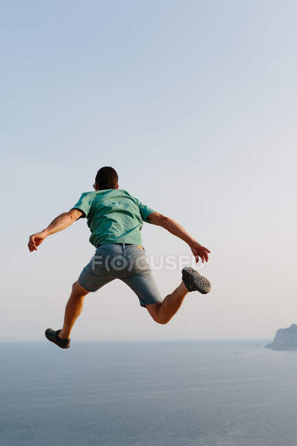 Low angle back view of unrecognizable male in casual clothes jumping high against blue sky and sea during summer evening — Stock Photo