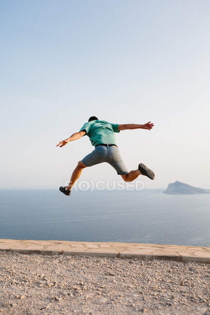 Vista posteriore ad angolo basso di un maschio irriconoscibile in abiti casual che salta in alto contro il cielo blu e il mare durante la sera d'estate — Foto stock