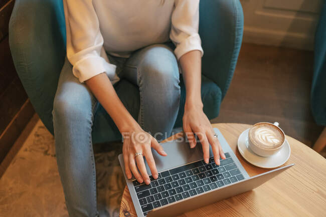 From above cropped unrecognizable young female freelancer in casual clothes having tasty hot drink while sitting at round wooden table and using netbook in light modern cafe — Stock Photo