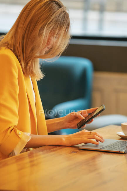 Mujer de negocios adulta en ropa formal mirando hacia otro lado mientras se sienta en la mesa de madera con el ordenador portátil en la cafetería contemporánea - foto de stock