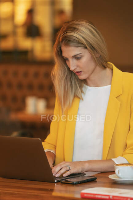 Взрослая деловая женщина в формальной одежде смотрит в сторону, сидя за деревянным столом с ноутбуком в современном кафе-магазине — стоковое фото